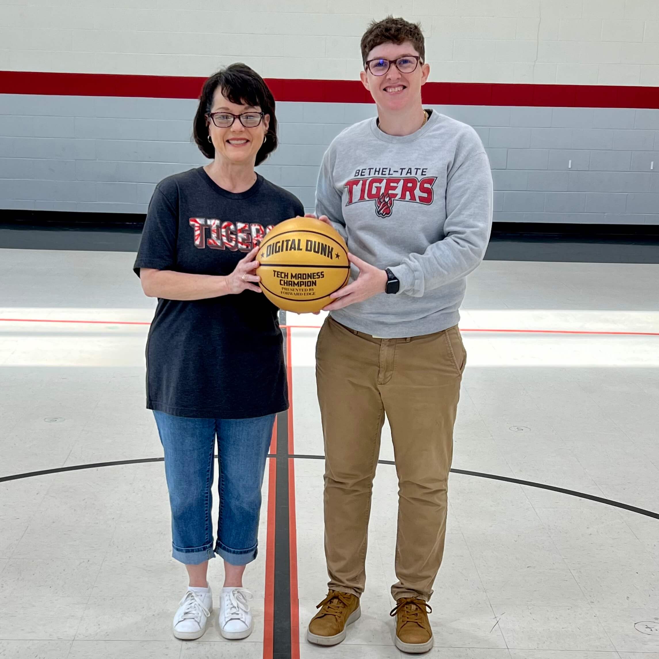 Ms. Patrick, a first-grade teacher, stands in a gymnasium alongside a Farmer Faubs, both smiling and holding a golden basketball labeled "Digital Dunk - Tech Madness Champion." She is wearing a black "Tigers" t-shirt, jeans, and white sneakers, while her colleague is dressed in a gray "Bethel-Tate Tigers" sweatshirt and khaki pants. The background features a basketball court with red and gray walls.